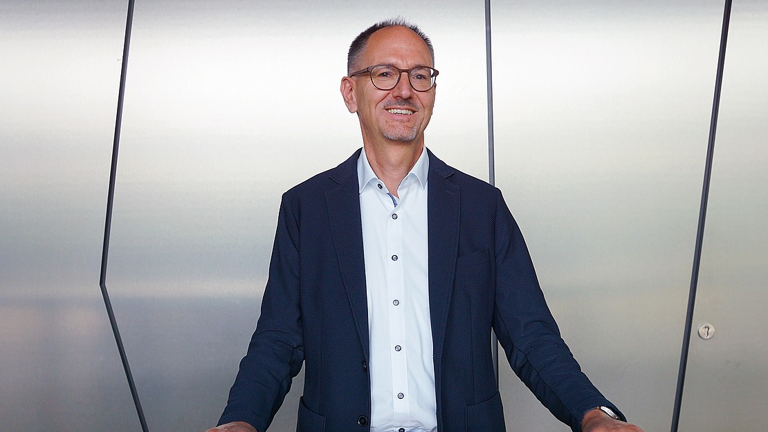 Ein Mann mit Brille, einem dunklen Blazer und einem hellen Hemd steht lächelnd vor einer metallischen Wand mit geometrischem Muster.