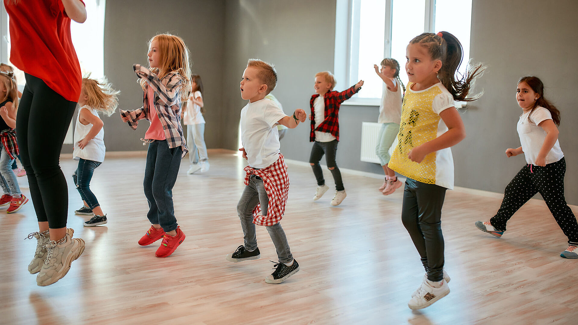 Eine Gruppe kleiner Kinder in Freizeitkleidung springt und tanzt gemeinsam in einem hellen, geräumigen Raum mit Holzboden und großen Fenstern. Ein Erwachsener leitet die Aktivität.