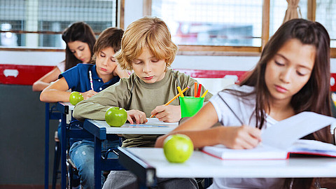 Vier Kinder sitzen an ihren Schreibtischen in einem Klassenzimmer und schreiben oder lesen. Auf jedem Schreibtisch liegen ein grüner Apfel und Schulmaterialien. Das Klassenzimmer hat große Fenster und wird von Tageslicht erhellt.