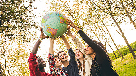 Fünf lächelnde junge Menschen stehen im Freien und strecken gemeinsam die Hände nach oben, um einen großen Globus zu halten; im Hintergrund sind Bäume und Sonnenlicht zu sehen.