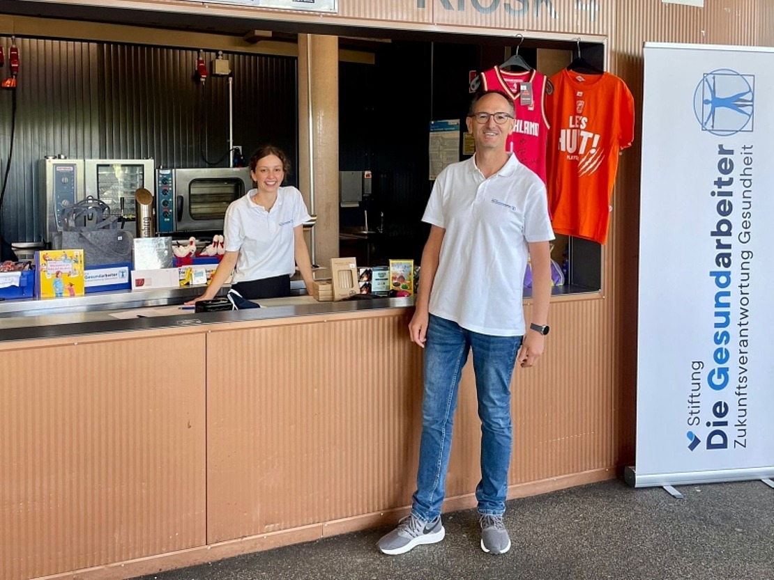 Zwei Personen in weißen Hemden stehen an einem Verkaufsstand mit Lebensmitteln und Sporttrikots. Auf einem vertikalen Banner rechts steht „Stiftung Die Gesundheitsarbeiter“. Beide lächeln in einer ungezwungenen Umgebung in einem Innenraum.