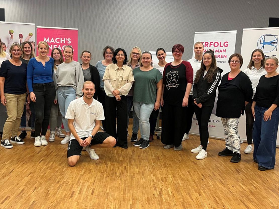 Eine Gruppe von 20 Personen, überwiegend Frauen, posiert in einem Raum auf einem Holzboden für ein Foto, im Hintergrund sind Transparente zu sehen. Ein Mann kniet vorne, während die übrigen stehen und in die Kamera lächeln.