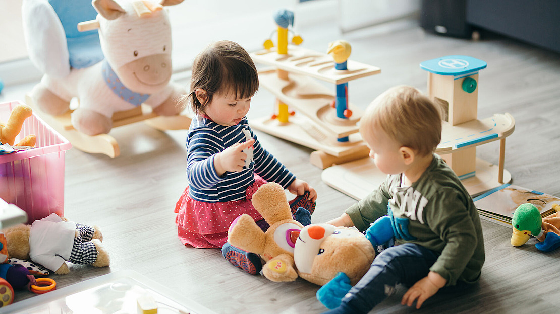 Zwei Kleinkinder sitzen auf dem Boden in einem Spielzimmer und spielen mit einem Plüschbären. Um sie herum liegen verschiedene Holz- und Plüschspielzeuge verstreut, und durch ein Fenster fällt Sonnenlicht herein.