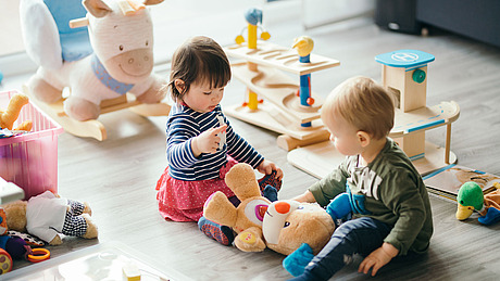 Zwei Kleinkinder sitzen auf dem Boden in einem Spielzimmer und spielen mit einem Plüschbären. Um sie herum liegen verschiedene Holz- und Plüschspielzeuge verstreut, und durch ein Fenster fällt Sonnenlicht herein.