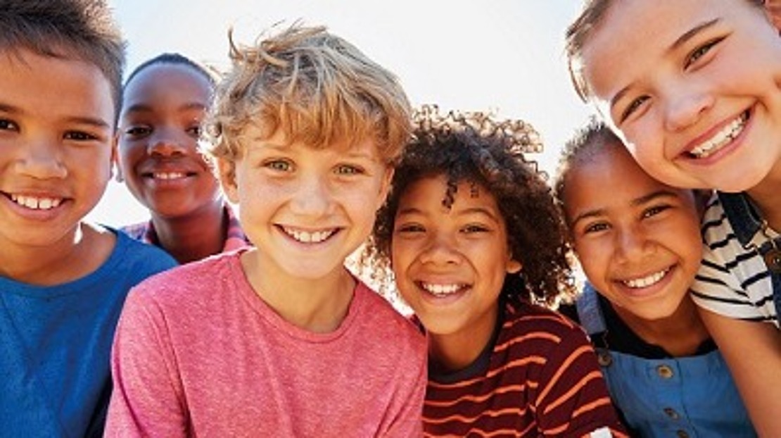 Eine Gruppe von sechs lächelnden Kindern steht im Freien dicht beieinander und blickt in die Kamera. Hinter ihnen scheint die Sonne hell und sorgt für eine fröhliche und freundliche Atmosphäre.