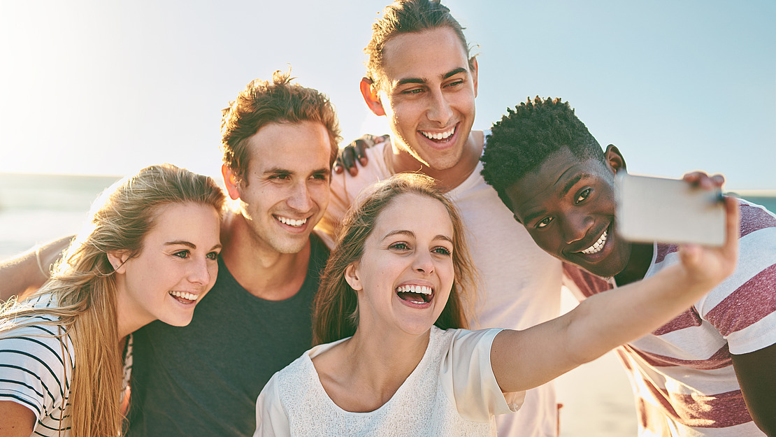 Fünf junge Erwachsene, die an einem sonnigen Tag im Freien lächeln und gemeinsam ein Selfie machen, vor dem Hintergrund eines blauen Himmels.