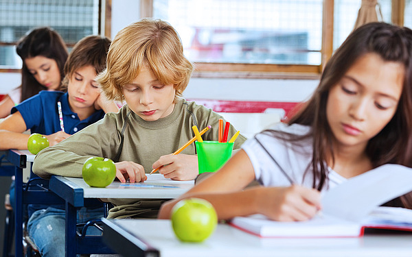Vier Kinder sitzen an ihren Schreibtischen in einem Klassenzimmer und schreiben oder malen. Auf jedem Schreibtisch liegen ein grüner Apfel und Schreibutensilien. Das Klassenzimmer hat Fenster und rot-weiße Wände. Die Schüler scheinen sich ganz auf ihre Arbeit zu konzentrieren.