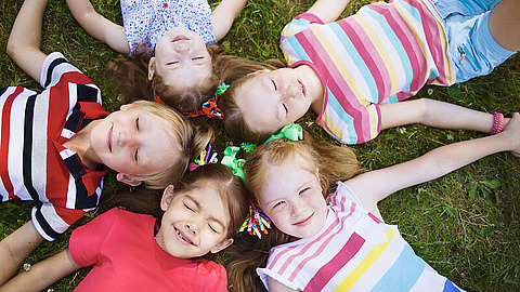 Fünf Kinder liegen an einem sonnigen Tag im Kreis auf dem Rasen, die Köpfe aneinander gelehnt, tragen bunte Kleidung und lächeln in die Kamera.